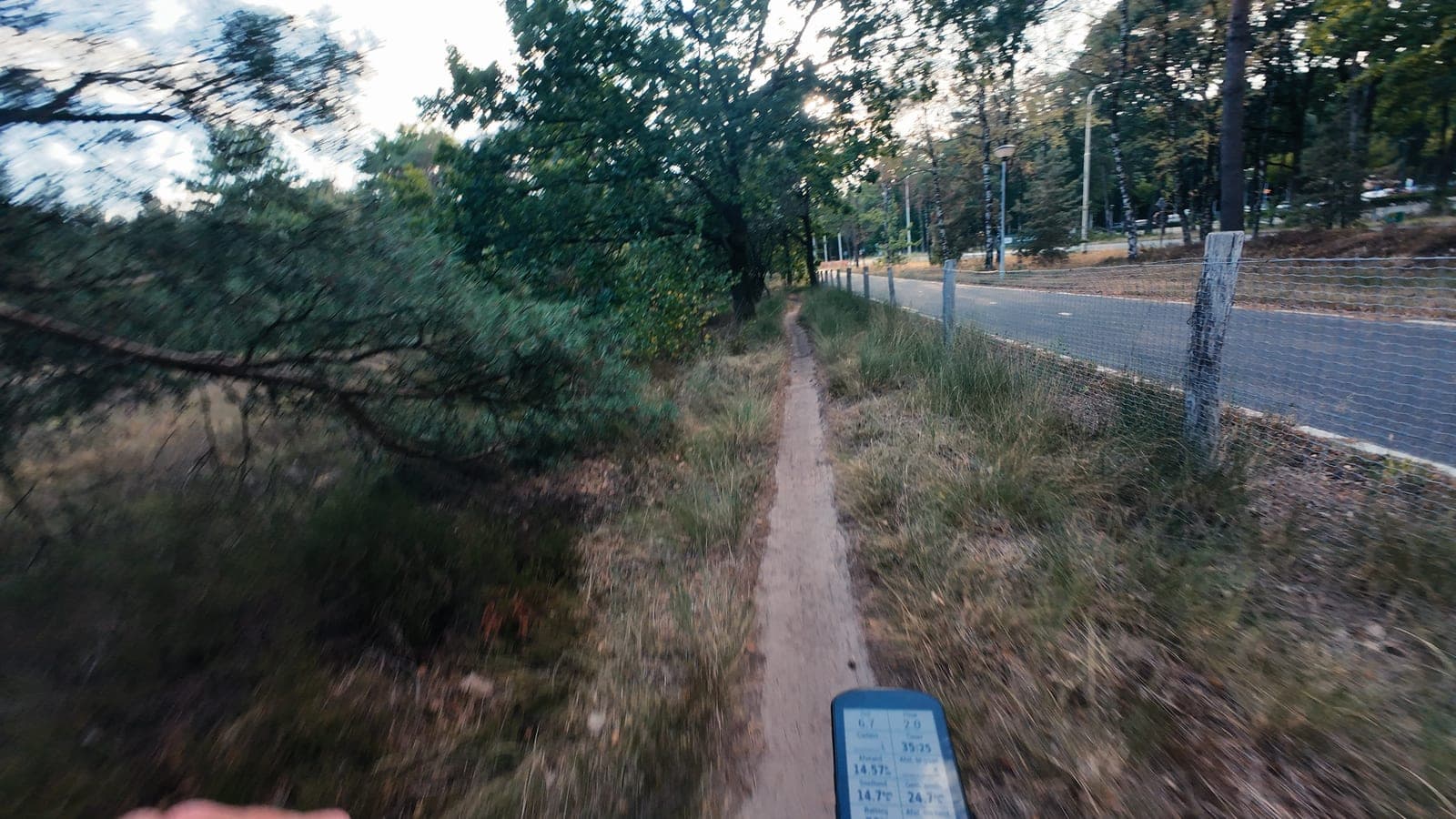 POV over het stuur met een hand aan het handvat en een fietscomputer, zandpad naast een wildraster