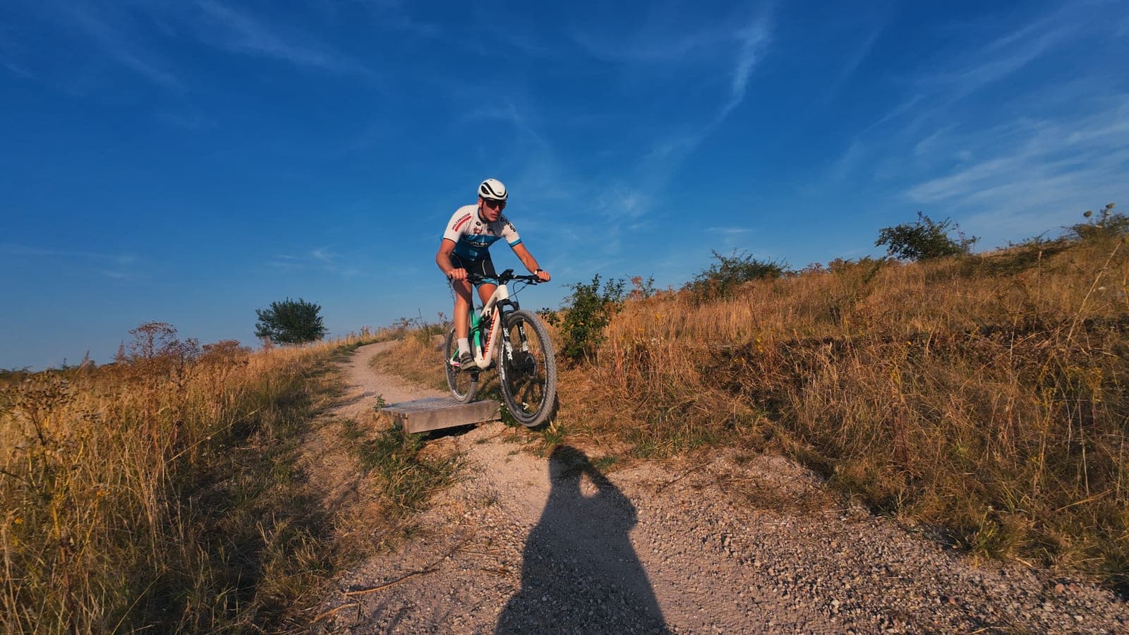 Mountainbiker rijdt van een houten obstakel af op een zandpad onder een blauwe lucht